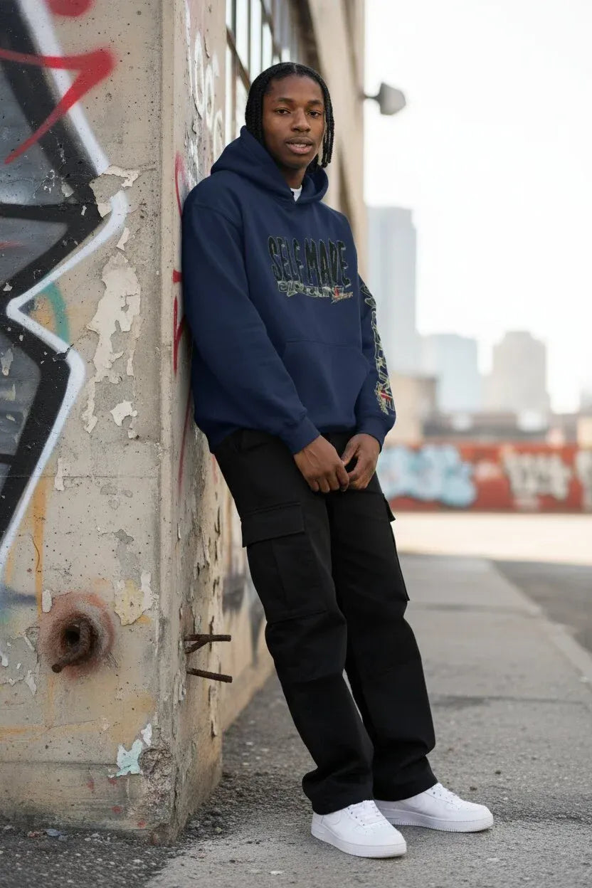 A man wearing a navy blue 'SELF MADE' hoodie, black cargo pants, and white sneakers stands leaning against a graffiti-covered concrete wall in an urban outdoor setting.
