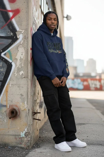 A man wearing a navy blue 'SELF MADE' hoodie, black cargo pants, and white sneakers stands leaning against a graffiti-covered concrete wall in an urban outdoor setting.