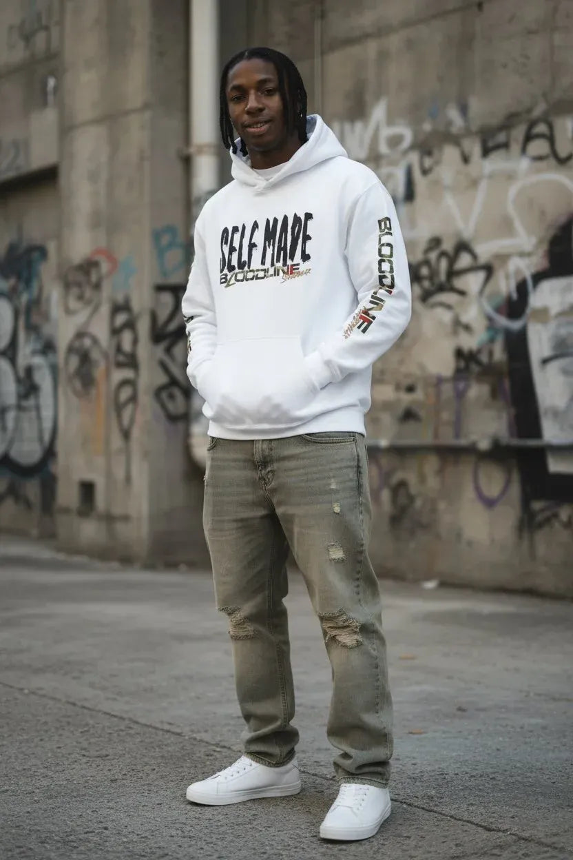 A man wearing a white pullover hoodie with 'SELF MADE ' printed on the front, paired with distressed olive green jeans and white sneakers, standing in front of a graffiti-covered concrete wall.