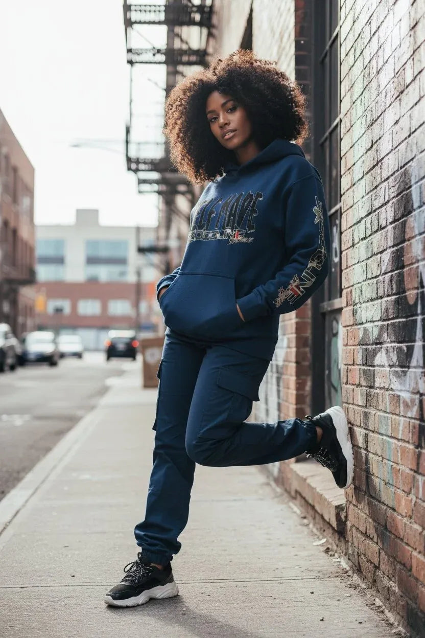 A woman with curly hair models a matching navy blue SELF MADE hoodie and jogger sweatpants set on an urban sidewalk next to a brick wall.
