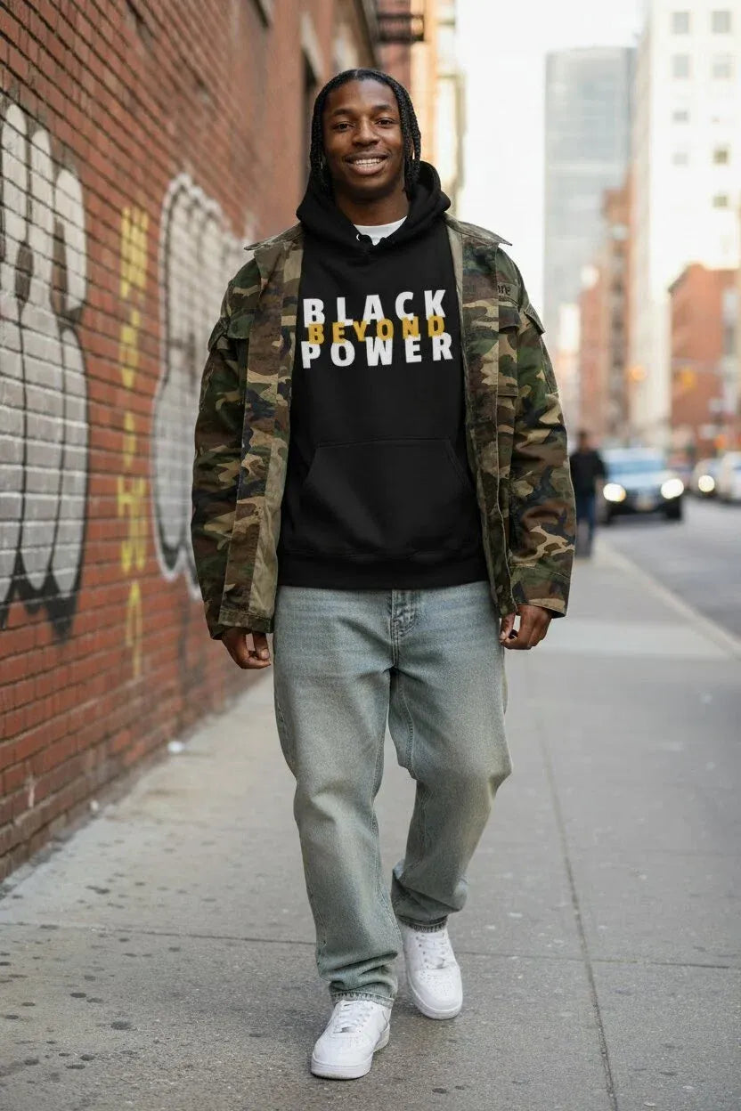 A man walking on a city sidewalk wearing a black hoodie with 'BLACK POWER' printed in yellow and white, layered under a camouflage jacket, paired with light gray jeans and white sneakers.