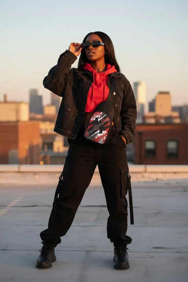 A woman stands on a rooftop wearing a black denim jacket, black cargo pants, a bright red hoodie, black boots, and a red, black, and white patterned crossbody bag. The background shows a blurred city skyline at sunset.