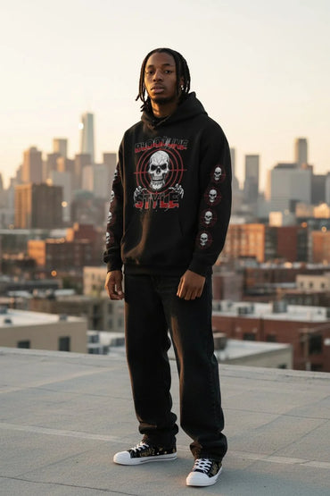 A man stands on a rooftop wearing a black graphic hoodie with a skull design and text, black sweatpants, and black-and-white sneakers, with a blurred city skyline in the background.