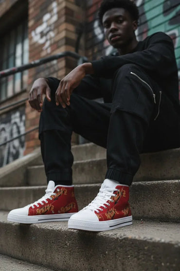 A man sitting on concrete stairs wearing black sweatpants and red high-top sneakers with gold graphic patterns and white soles, set against an urban brick wall background.
