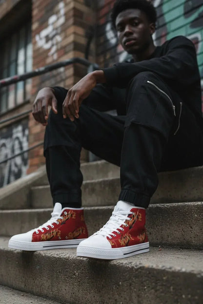 A man sitting on concrete stairs wearing black sweatpants and red high-top sneakers with gold graphic patterns and white soles, set against an urban brick wall background.