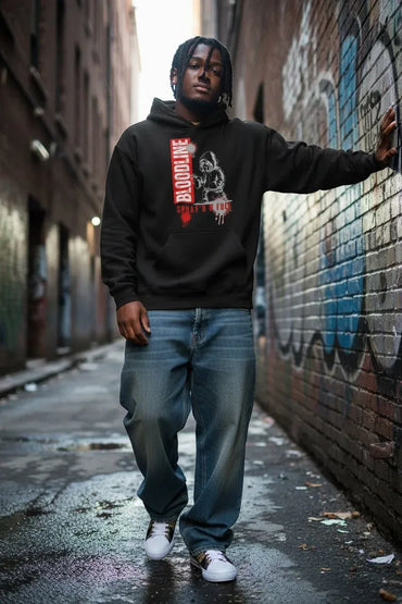 A man wearing a black graphic hoodie with a red and white design, blue jeans, and white sneakers, standing in a wet urban alleyway next to a graffiti-covered brick wall.