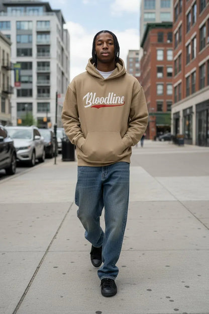 A man walking on a city sidewalk wearing a beige hoodie with 'Bloodline' printed across the chest in white and red, paired with blue jeans and black sneakers.
