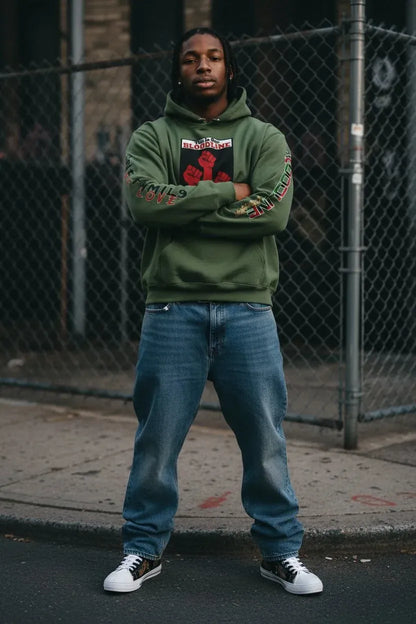 A man stands on a city sidewalk in front of a chain-link fence, wearing an olive green hoodie with a red and black graphic on the chest and 'LOVE' printed down both sleeves, paired with blue jeans and white sneakers.
