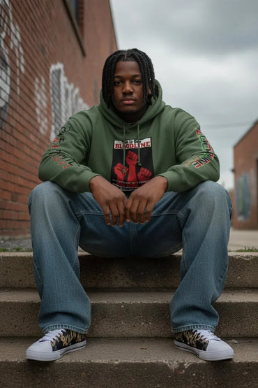 A man sits on concrete steps wearing an olive green graphic hoodie, blue jeans, and white sneakers with leopard print details. The background features a brick wall and an overcast sky.