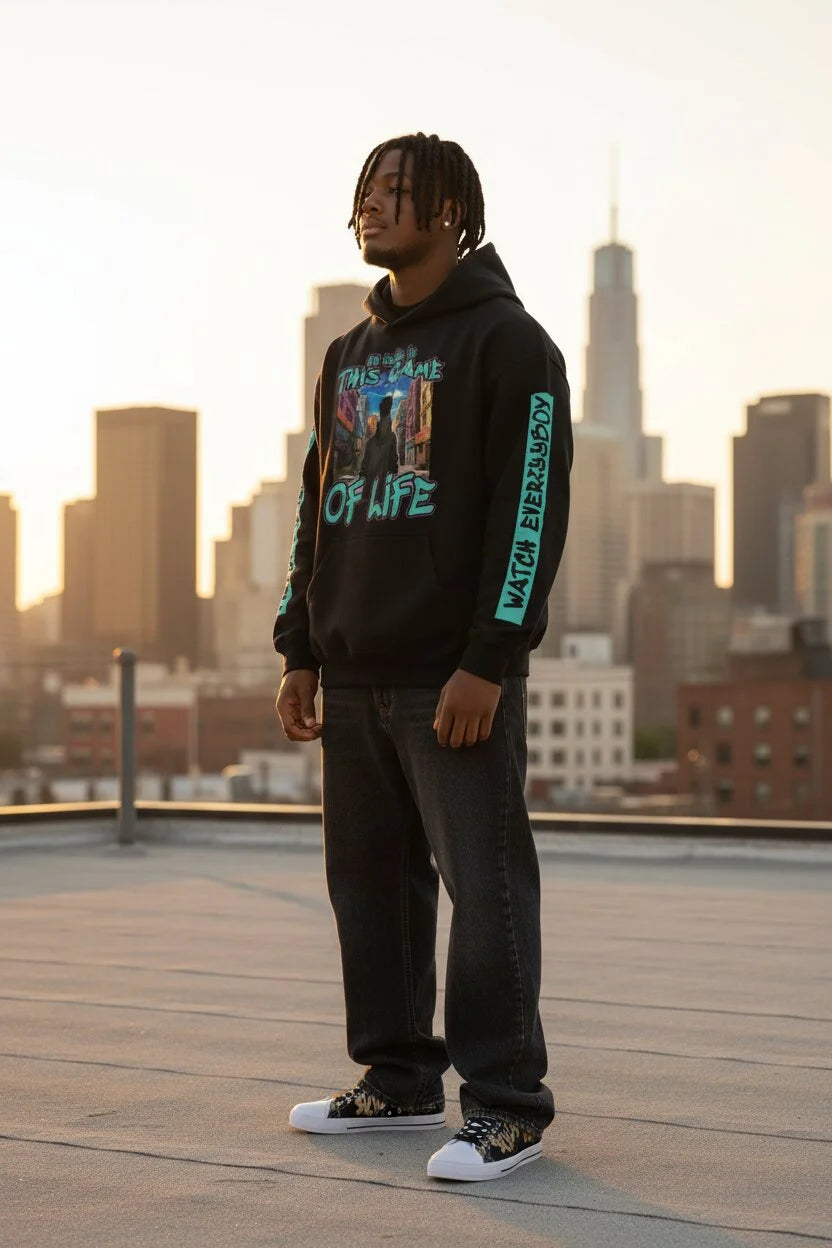 A man stands on a rooftop wearing a black graphic hoodie with turquoise and multicolor print, dark jeans, and black-and-white sneakers, with a city skyline in the background at sunset.