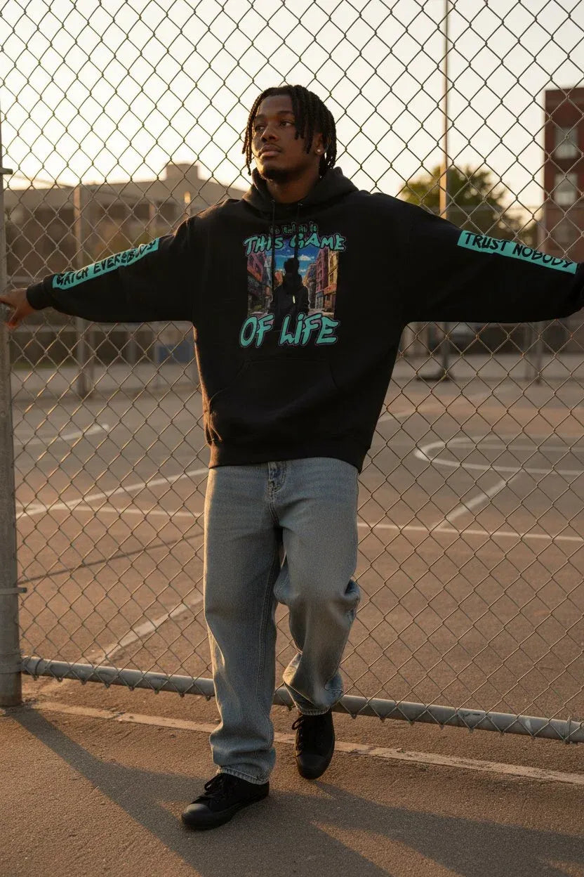 A man wearing a black graphic hoodie with 'TRUST NOBODY OF LIFE' printed on the front and 'TRUST NOBODY' on the sleeves, paired with light blue jeans, standing in front of a chain-link fence at sunset.