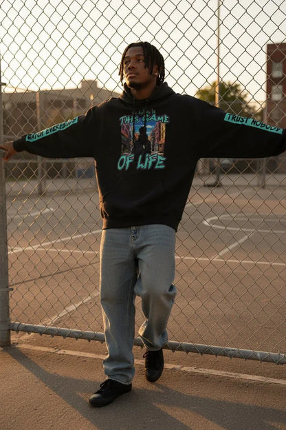A man wearing a black graphic hoodie with 'TRUST NOBODY OF LIFE' printed on the front and 'TRUST NOBODY' on the sleeves, paired with light blue jeans, standing in front of a chain-link fence at sunset.