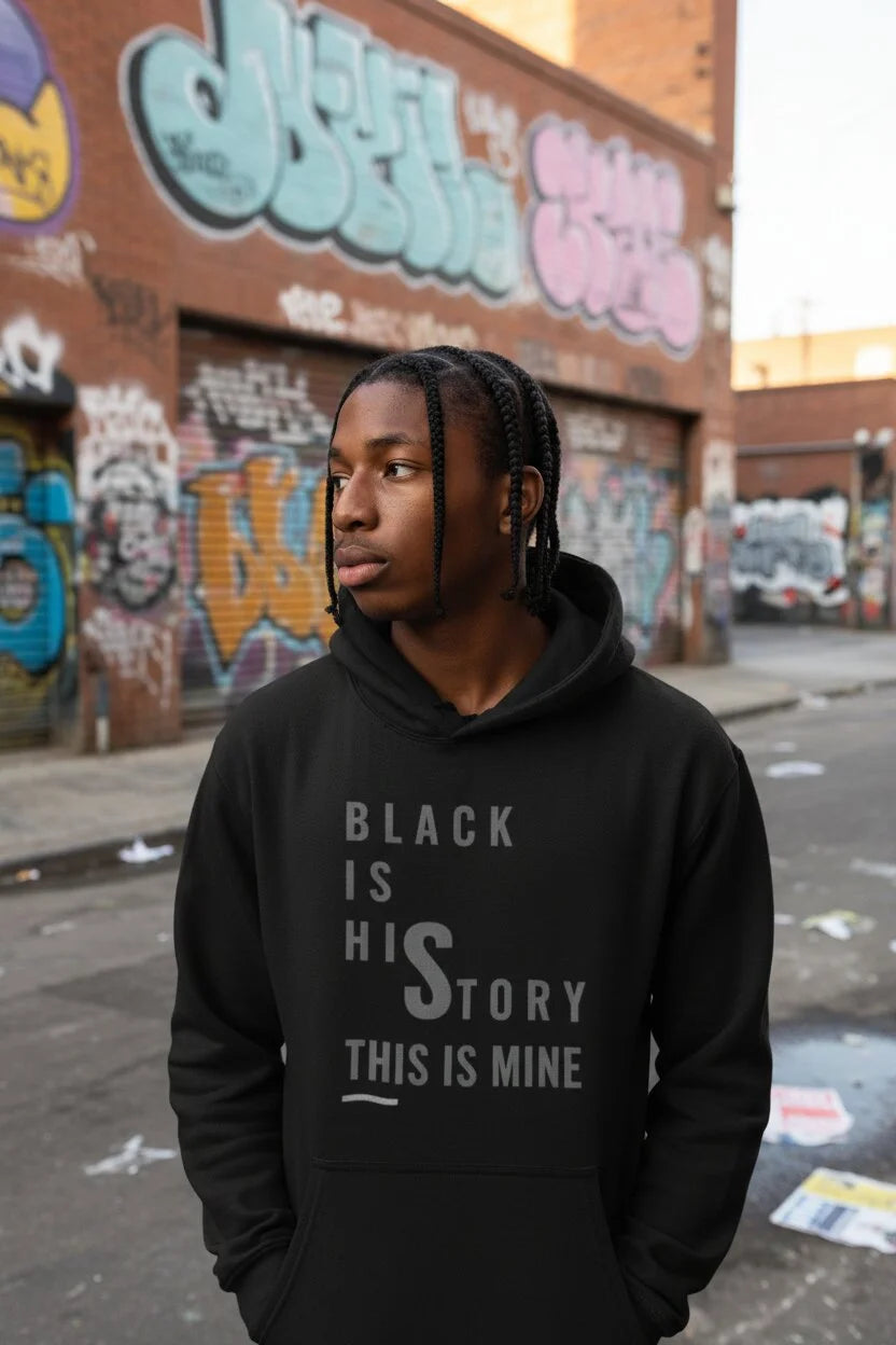 A man wearing a black hoodie with the text 'BLACK IS HIS STORY THIS IS MINE' printed in gray on the front, standing in an urban street with graffiti-covered brick walls in the background.