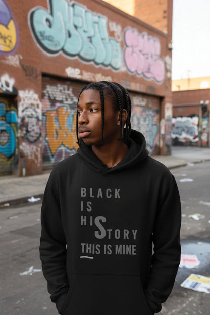 A man wearing a black hoodie with the text 'BLACK IS HIS STORY THIS IS MINE' printed in gray on the front, standing in an urban street with graffiti-covered brick walls in the background.