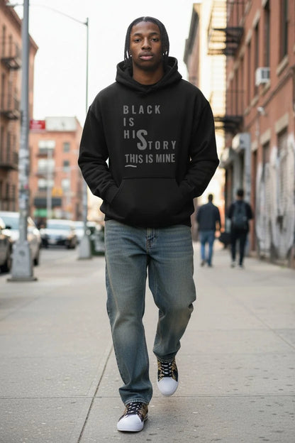 A man walking on an urban sidewalk wearing a black pullover hoodie with the text 'BLACK IS HIS STORY THIS IS MINE' printed in white on the front, paired with blue jeans and white sneakers.