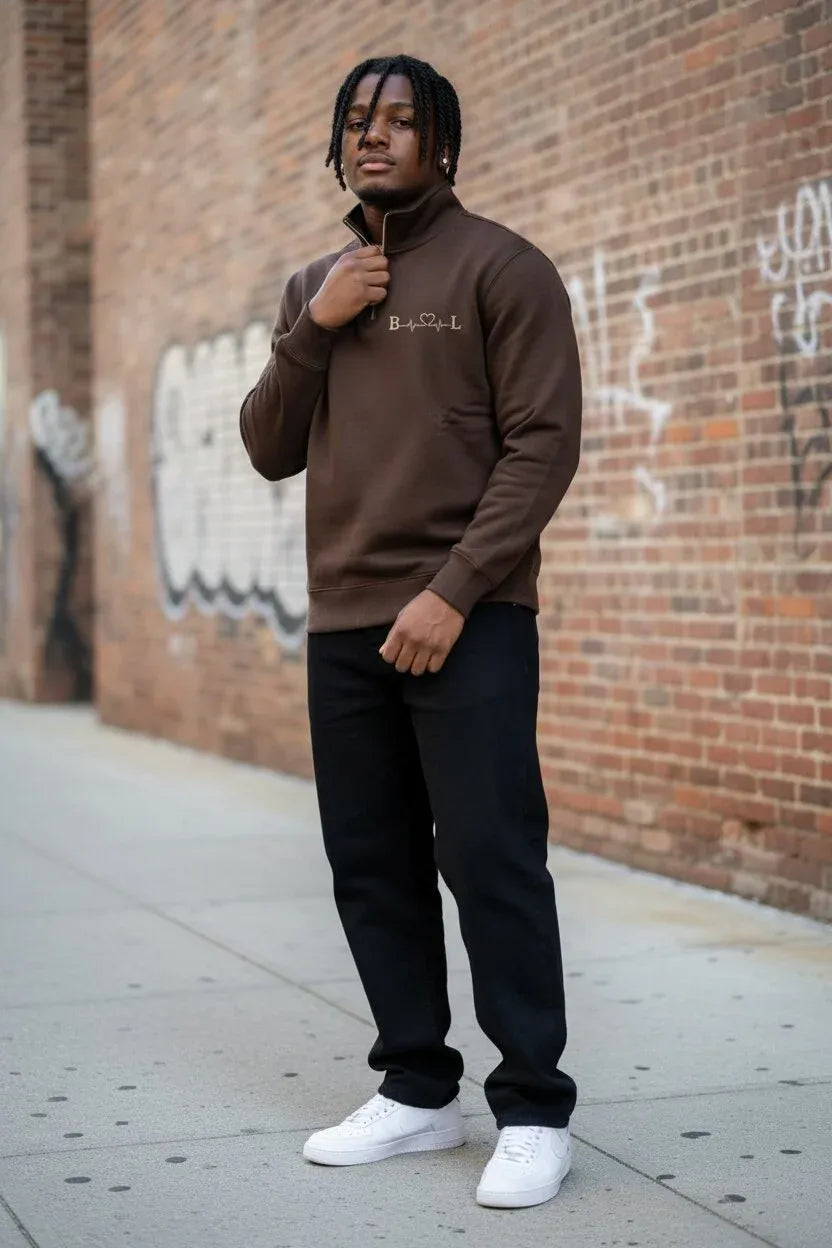 A man wearing a brown quarter-zip sweatshirt with a small white embroidered logo on the chest, black pants, and white sneakers, standing on a city sidewalk against a brick wall with graffiti.
