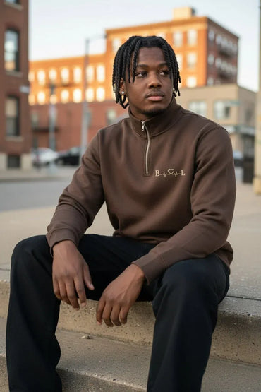 A man with dreadlocks sits on a concrete curb in an urban setting, wearing a brown long-sleeve quarter-zip sweatshirt with 'B. Soul' embroidered on the chest, paired with black sweatpants.