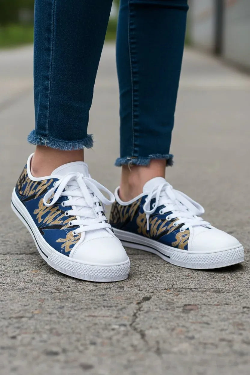 White low-top sneakers with a navy blue and gold abstract pattern on the sides, worn with frayed blue jeans on a concrete surface.