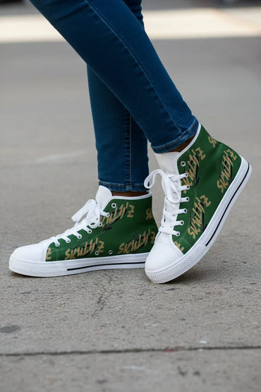 A person wearing green high-top sneakers with a gold script pattern and white laces, paired with blue jeans, standing on a gray concrete surface.