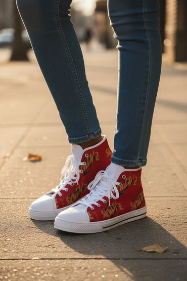 A person wearing red high-top sneakers with white rubber toe caps and white laces, standing on a sunlit sidewalk. The sides of the shoes are covered in a repeating gold script pattern.