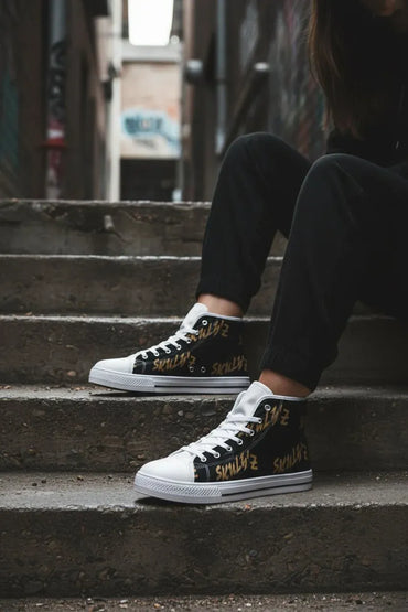 A person sitting on concrete steps wearing black high-top sneakers with white rubber toe caps and sides, white laces, and gold graffiti-style text reading 'I LOVE NY'.