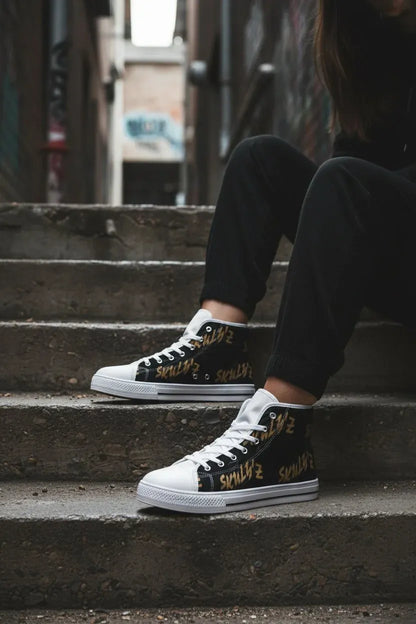 A person sitting on concrete steps wearing black high-top sneakers with white rubber toe caps and sides, white laces, and gold graffiti-style text reading 'I LOVE NY'.